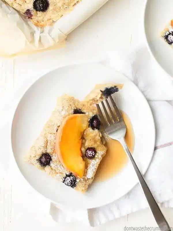 A plate with a slice of sheet pancakes topped with sliced peaches and maple syrup.