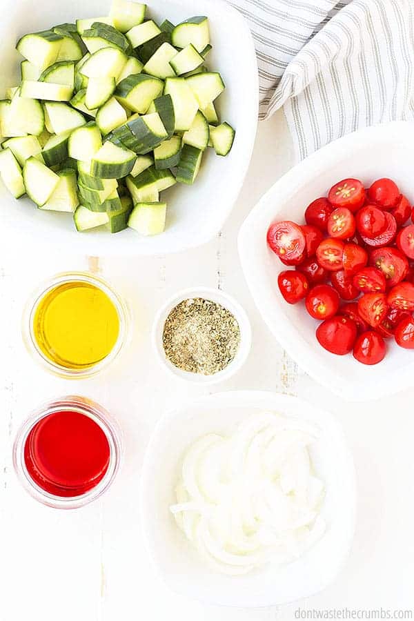 Sliced cucumber in a bowl, sliced cherry tomatoes in a bowl, sliced onions in a bowl, and three small glass jars with ingredients to make a dressing.