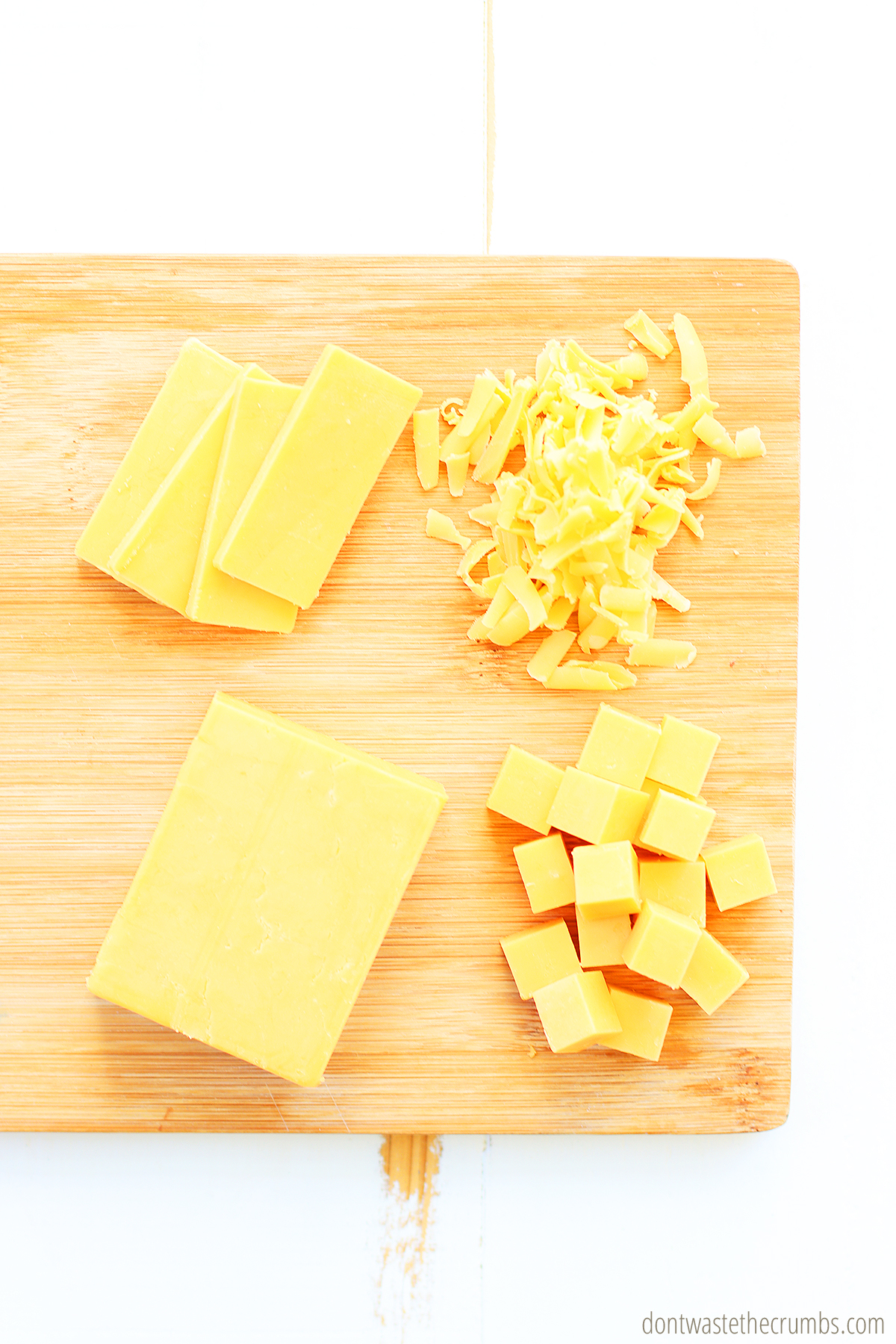 Sliced cheese, shredded cheese, a block of cheese, and cubes of cheese on a wood cutting board.