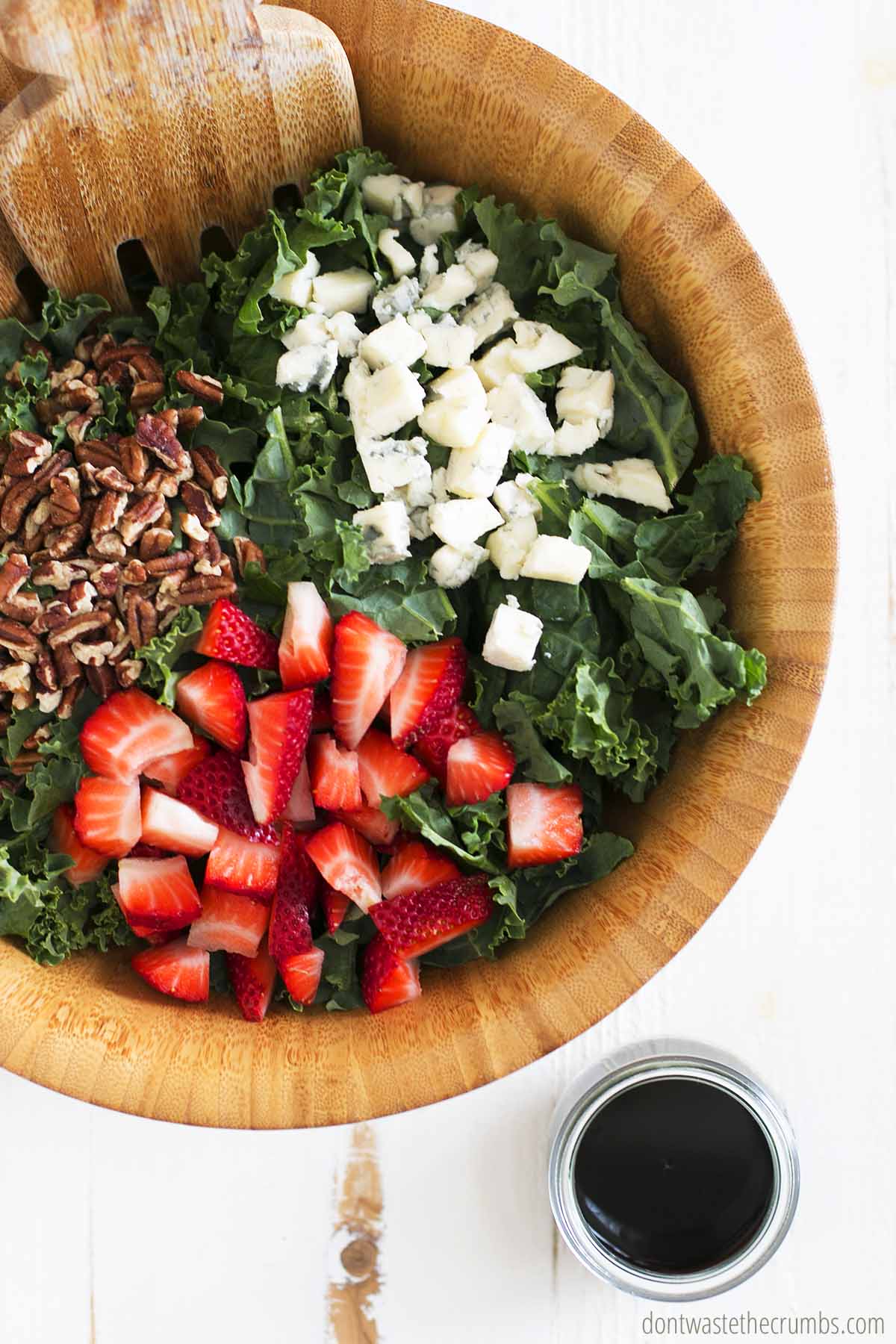 Chopped kale, strawberries, pecans, blue cheese in a wooden salad bowl ready to be tossed with balsamic vinaigrette dressing. 