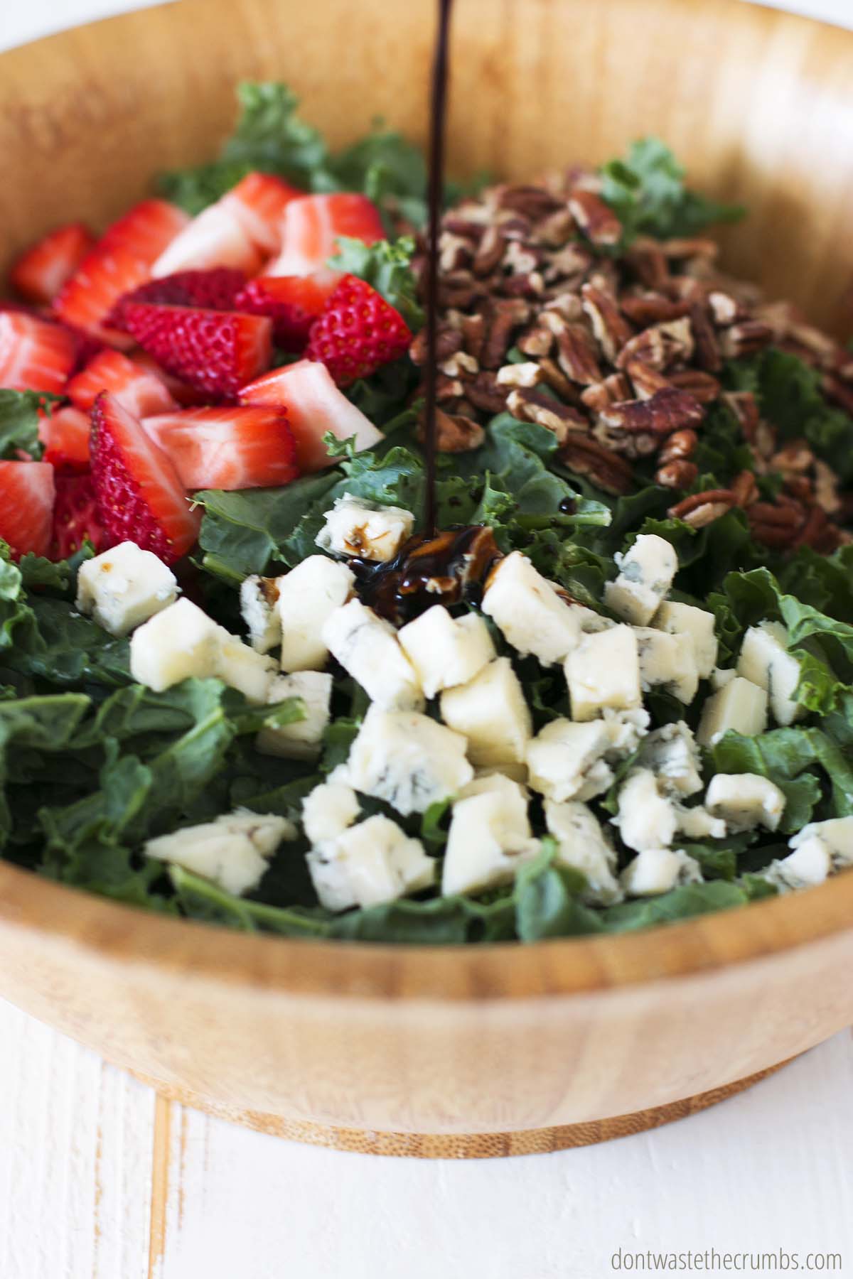 Pouring balsamic vinaigrette onto a kale salad recipe with strawberries, pecans and blue cheese.