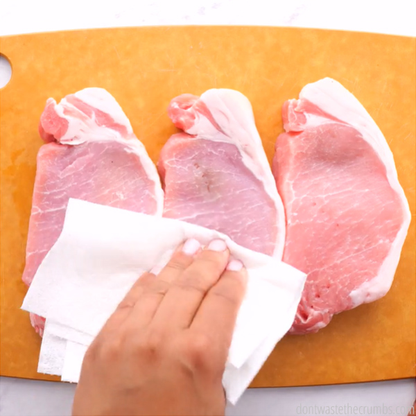 Three pork chops on a cutting board. A hand patting the meat dry with a paper towel.