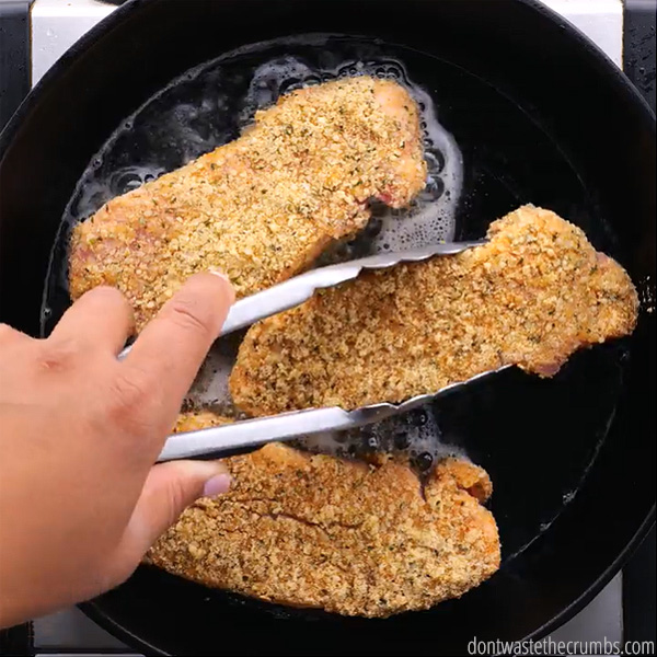 Fried pork chops in a cast iron skillet, cooking. A hand with tongs grabbing the pork chop.