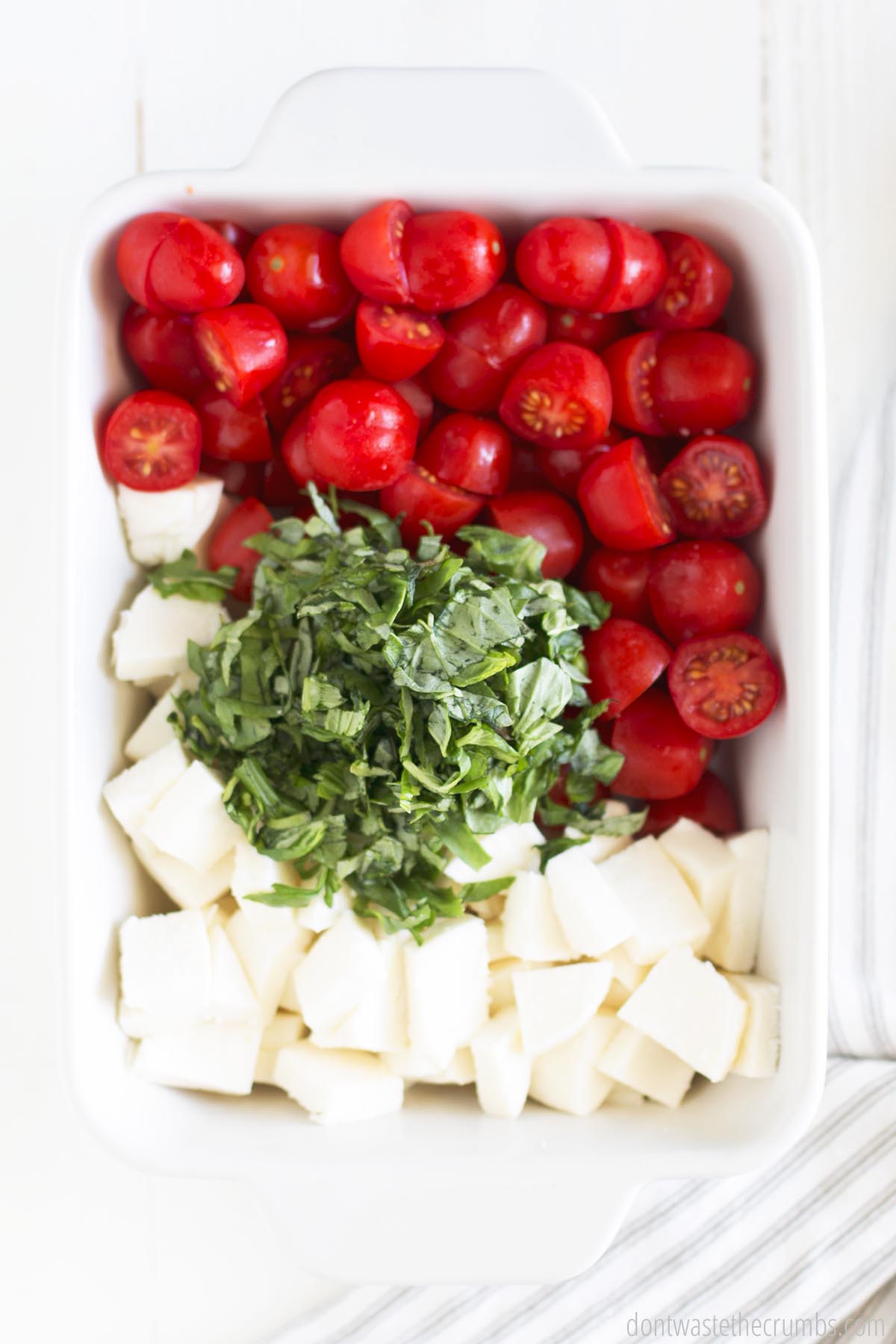 White baking dish with sliced cherry tomatoes, sliced mozzarella, and chopped basil.