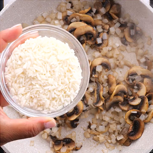 Chopped onions and mushroom in a frying pan. Hand holding a small bowl of rice over the frying pan.