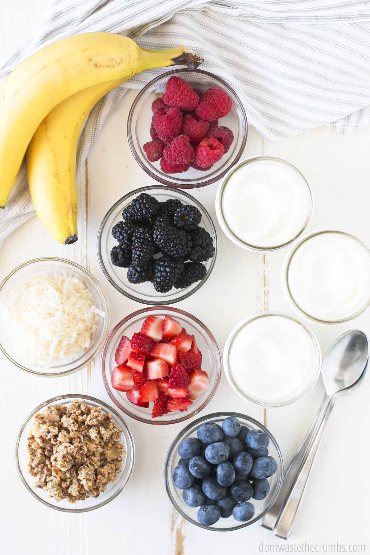 Two bananas next to ingredients for yogurt parfaits. Bowl of raspberries, bowl of black berries, bowl of coconut flakes, bowl of granola, bowl of sliced strawberries, bowl of blueberries, and three jars of yogurt. There are two spoons next to the jars.