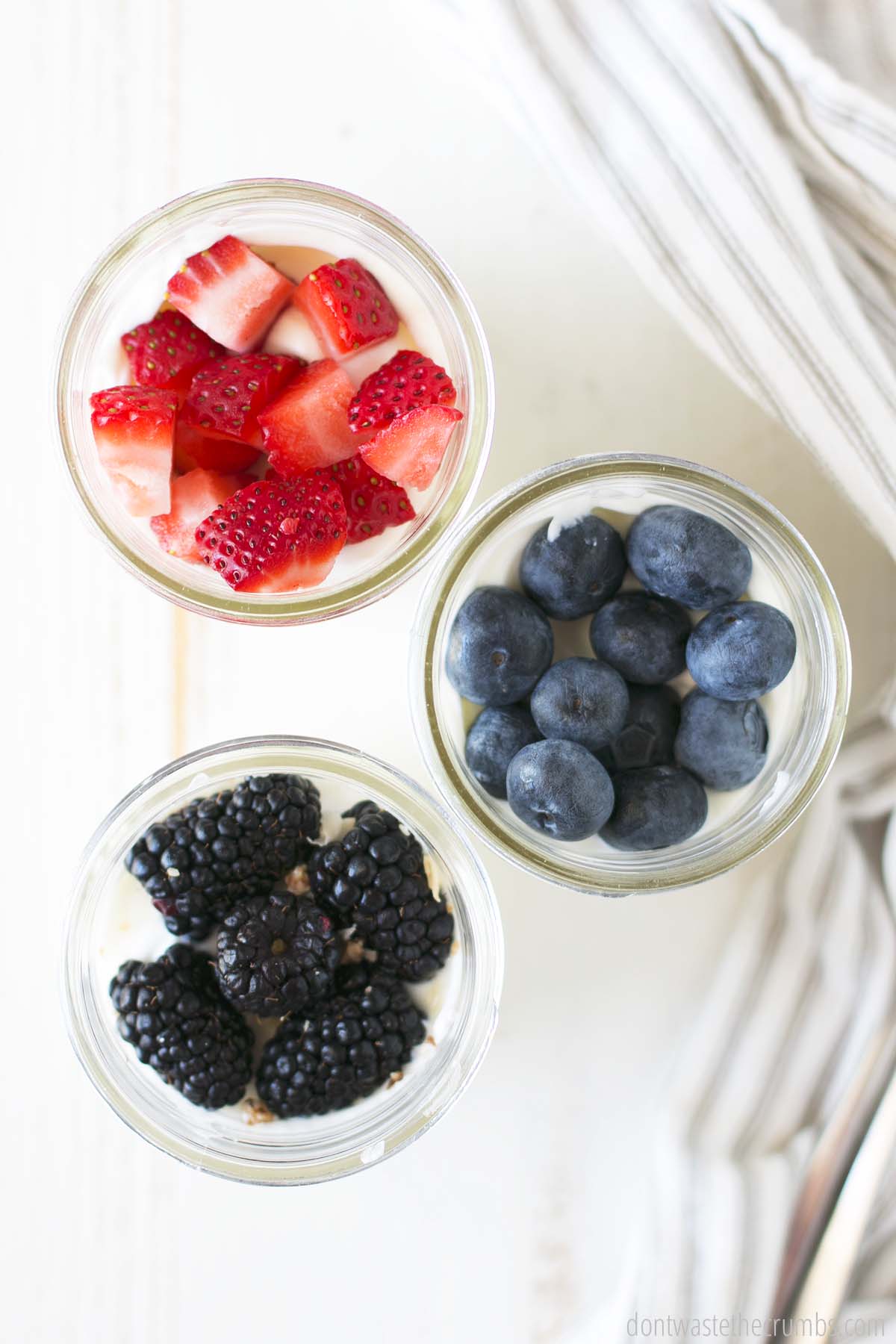 Overview of three yogurt parfaits. One has yogurt and sliced strawberries, the other has yogurt and blueberries, the other has yogurt and blackberries.