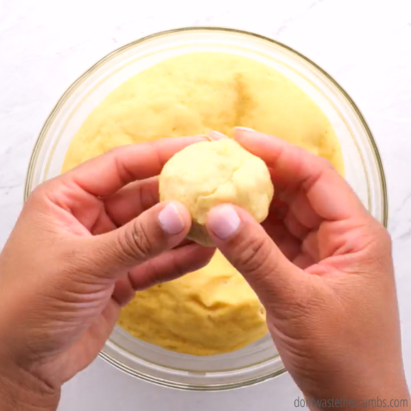 Hands rolling the dough into a ball shape for English muffins. There is a glass bowl with dough in the background.