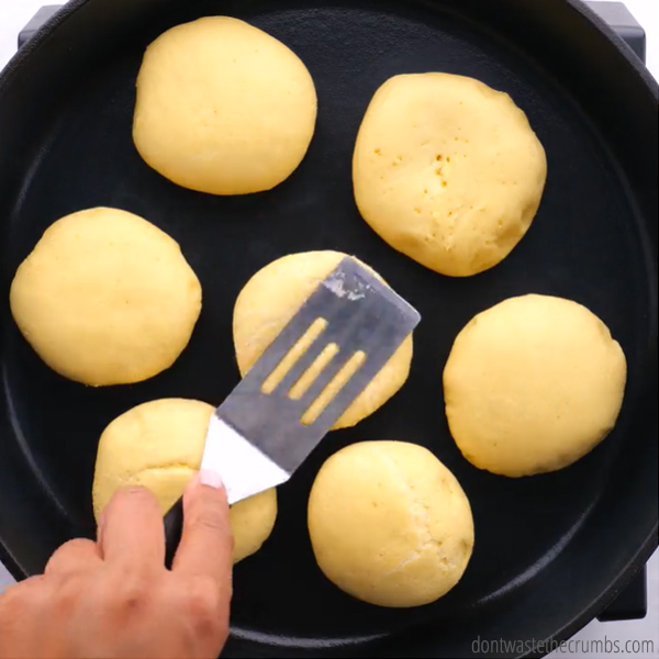 Seven dough balls on a cast iron skillet. Spatula is pressing on one ball.