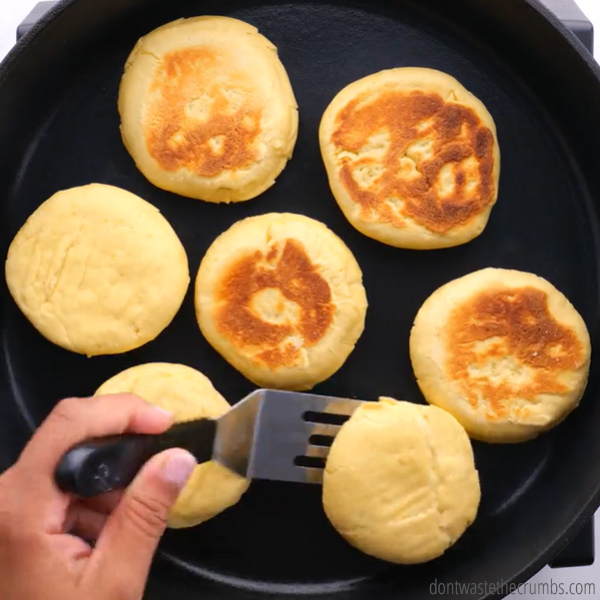 Seven English muffins on a cast iron skillet. A hand holding a spatula and flipping a muffin.
