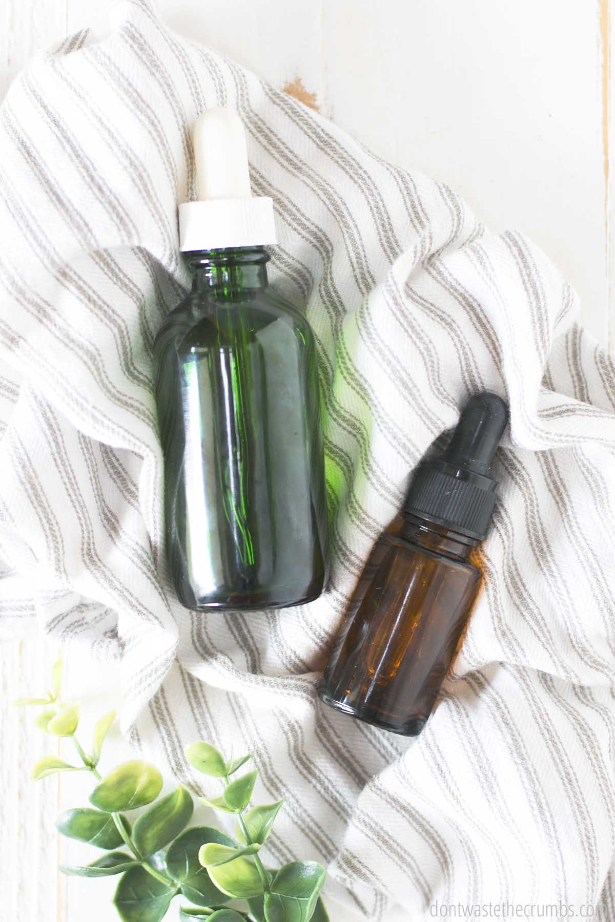 two dropper bottles laying on a white and blue cloth with greenery to the side.