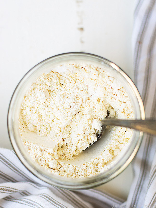 Overhead shot of white flour in a glass jar with a spoon. A blue and white striped towel in the background. 