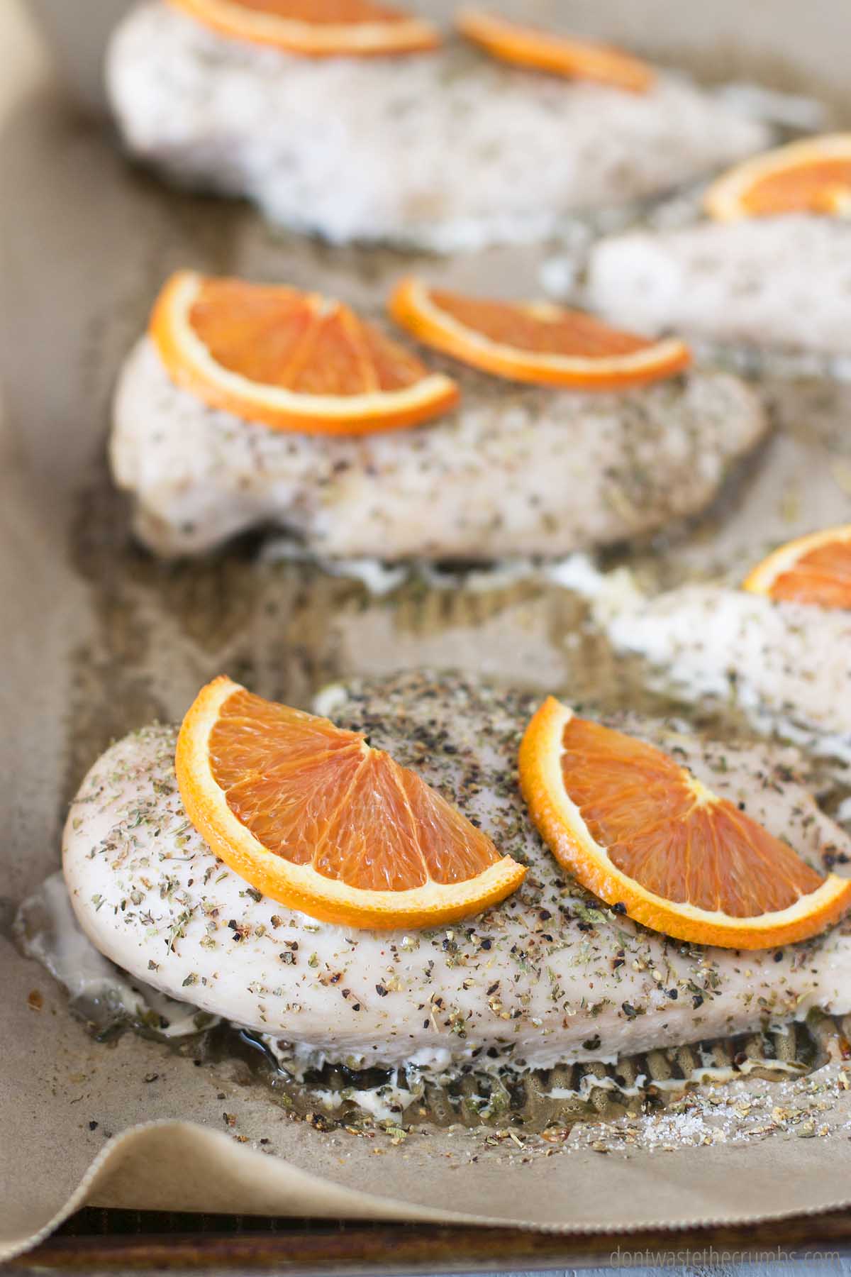 Cooked chicken breast seasoned with herbs and orange slices on baking sheet.