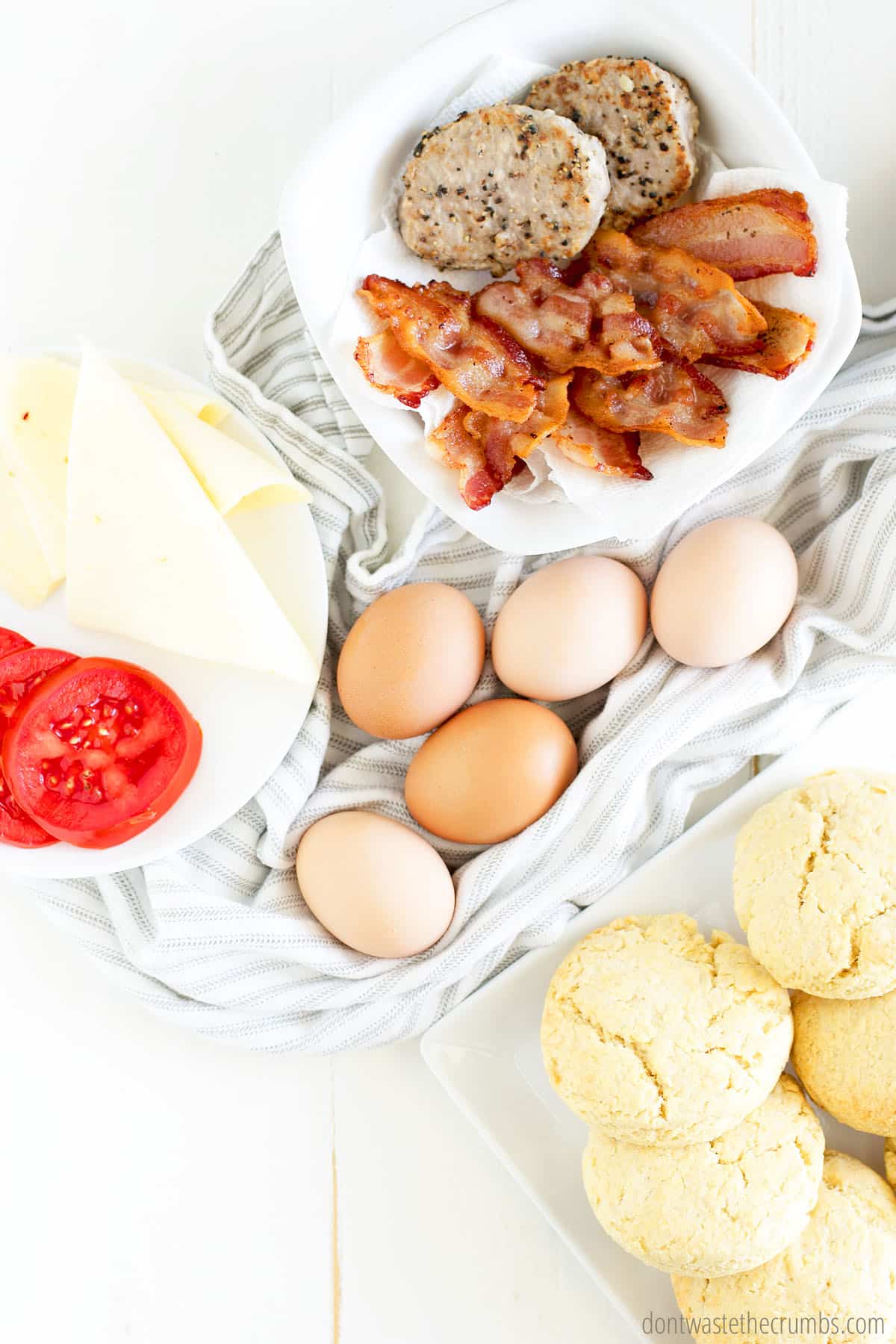 Ingredients for breakfast sandwich: sausage, bacon, cheese, sliced tomato, eggs, and biscuits.
