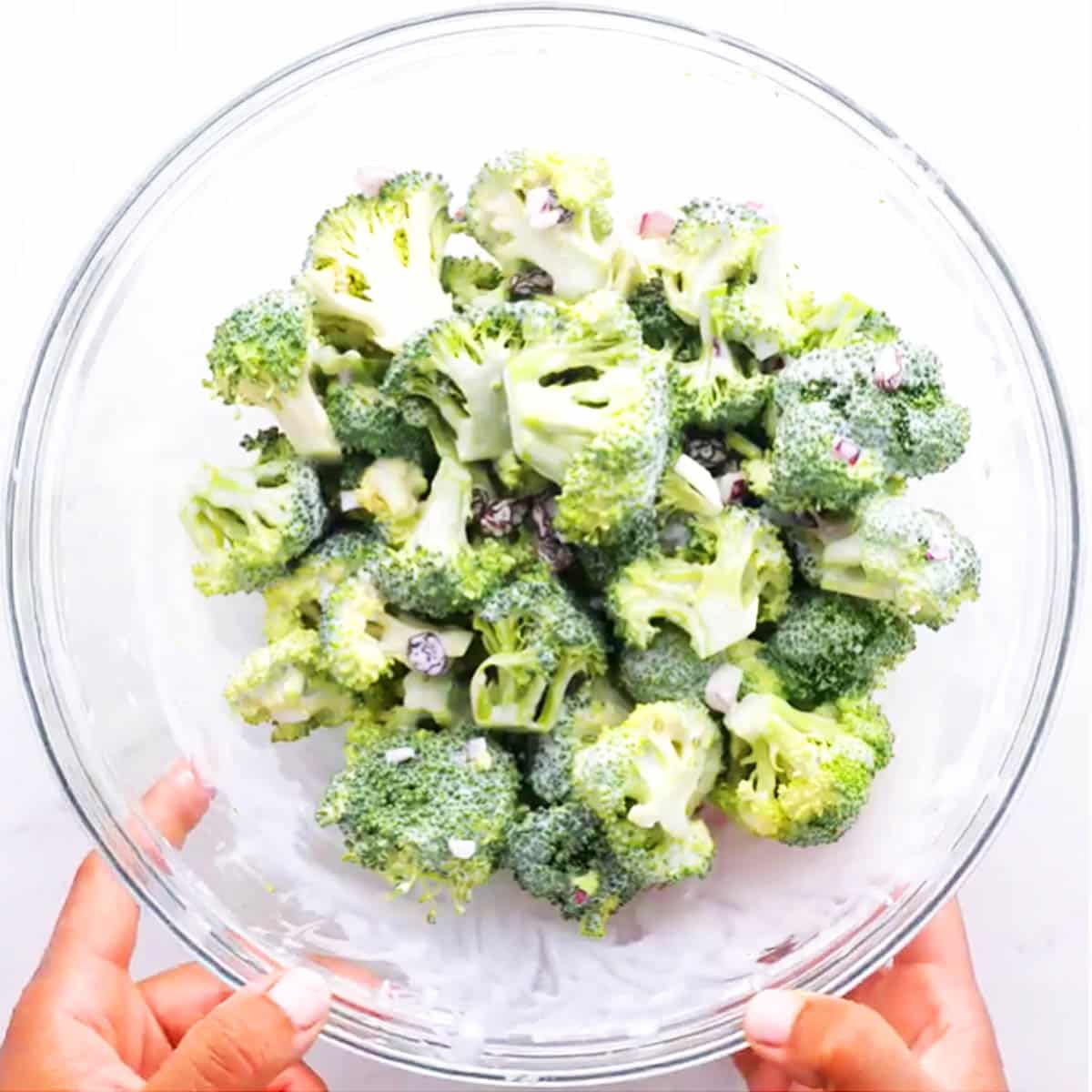 Two hands holding a large glass bowl of chopped broccoli and chopped red onion.