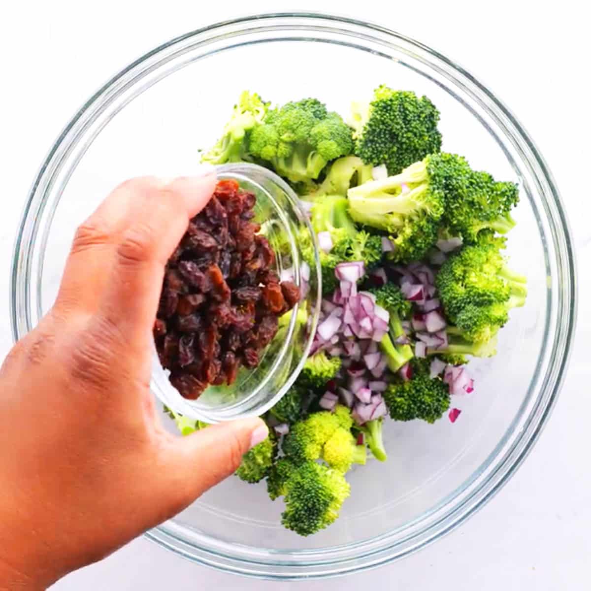 Hand pouring a small bowl of chopped bacon into the broccoli salad in a large glass bowl.