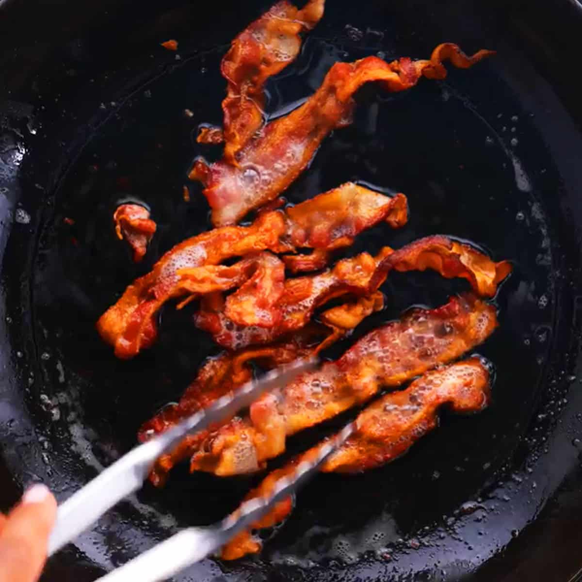 Hand holding tongs while adjusting bacon frying in a pan.