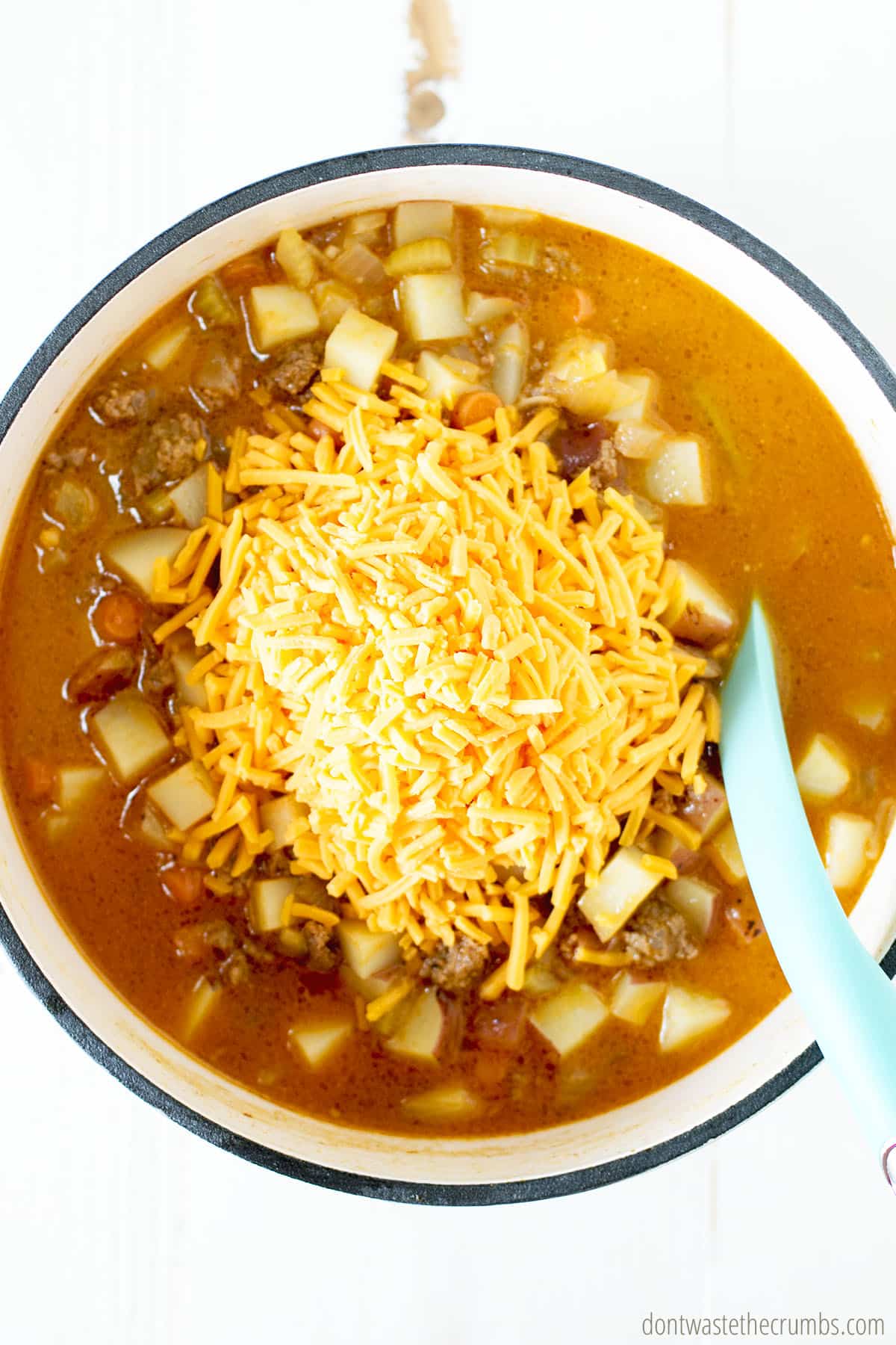Big pile of shredded cheese in a large put of cheeseburger soup. Large spoon stirring the soup.