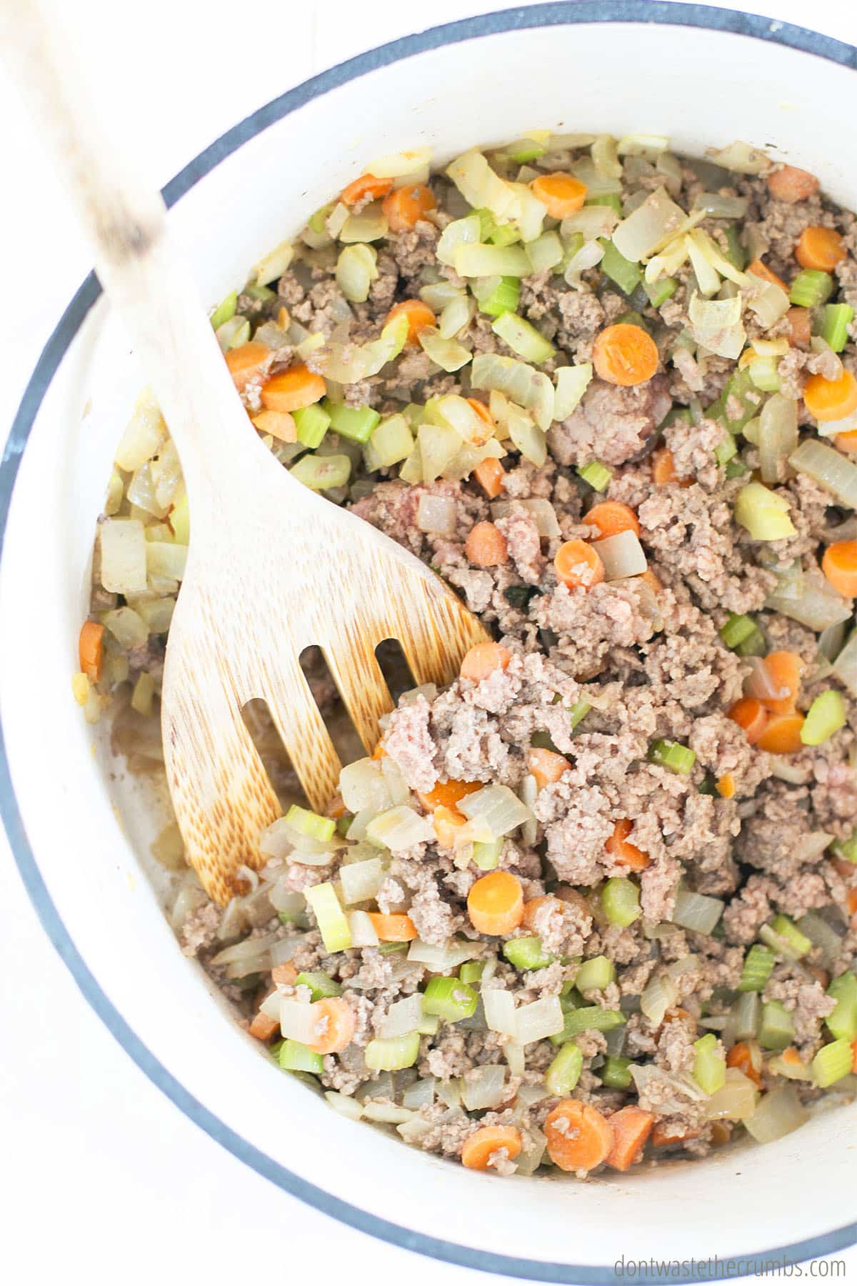 Large pot with ground beef and veggies, there is a wooden spatula stirring the mixture that makes cheeseburger soup.