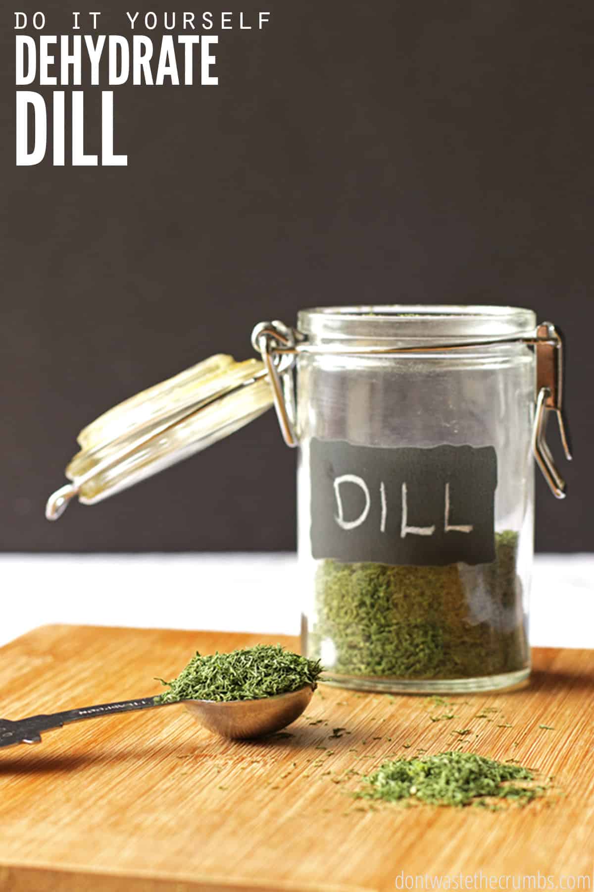 An open jar of dried dill, with a handwritten chalkboard label, sits on a wooden cutting board, with a measuring spoon full of dill to the left and a small pile of dried dill in front. Learn how to dry dill.