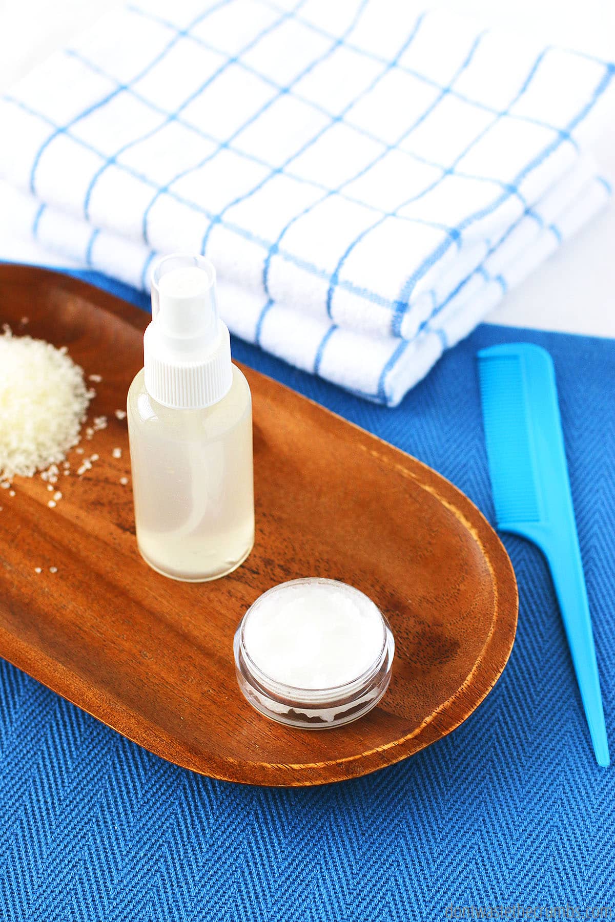 A wooden tray holds a container of a creamy conditioner, a plastic spray bottle with an apple cider rinse, and a pile of sea salt, with a comb to the right and a folded towel in the background.