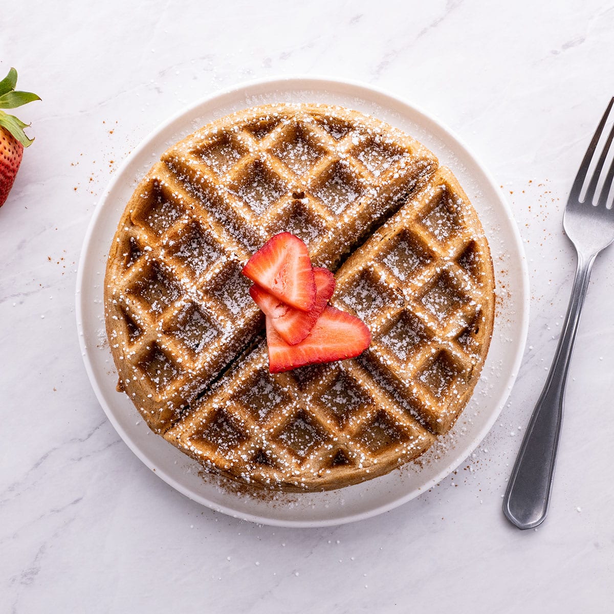oatmeal waffle sitting on a white plate with strawberries and powdered sugar on top.