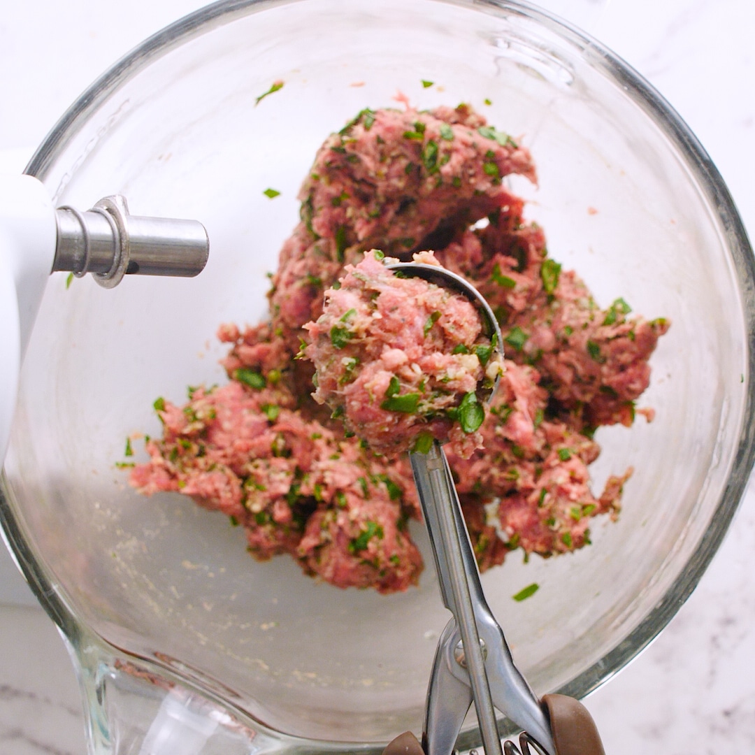 Raw meat for meatballs in a large mixing bowl. There is a full cookie scoop of the meat.