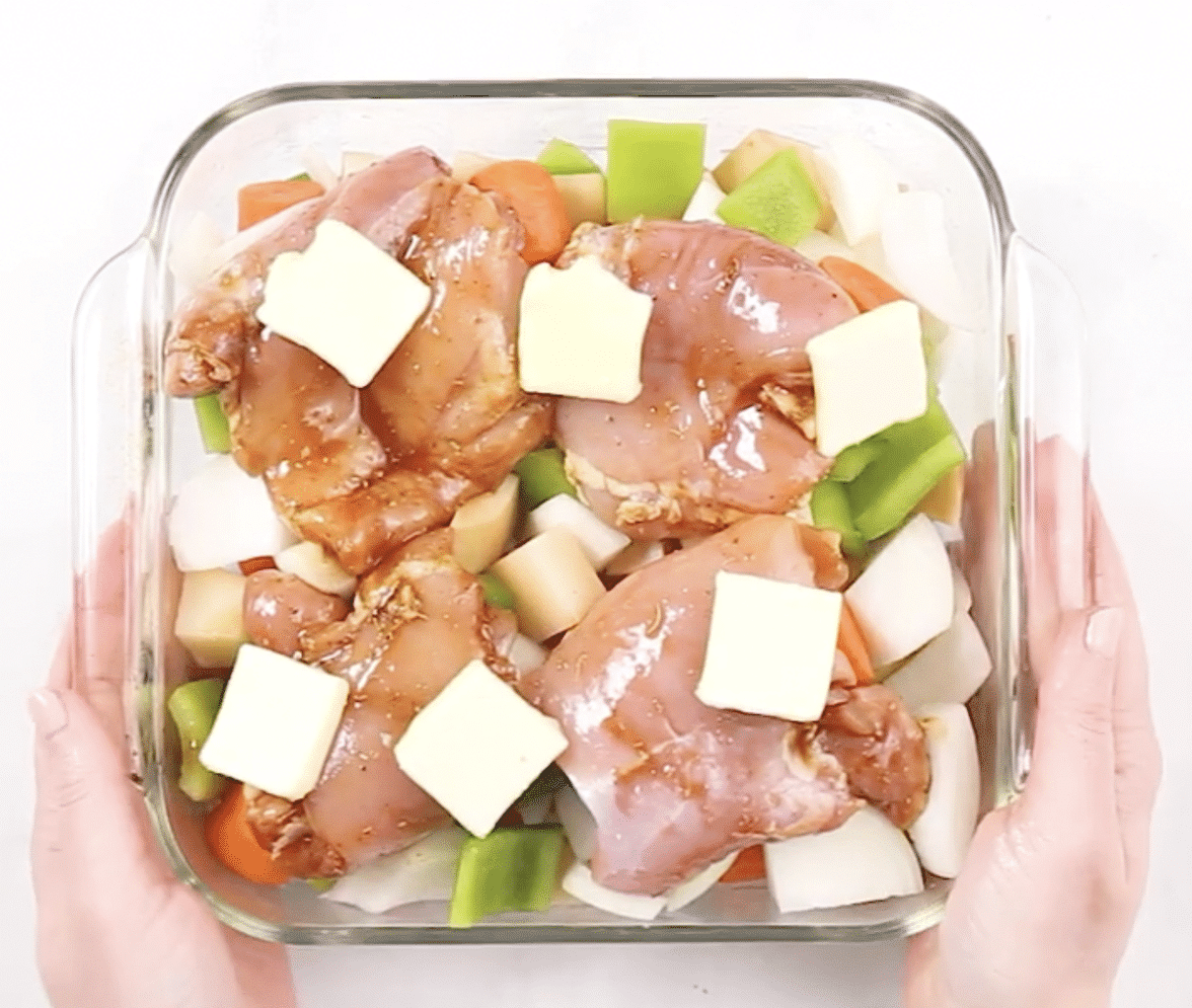 Hands holding a glass baking dish with veggies and raw chicken and slices of butter on the chicken