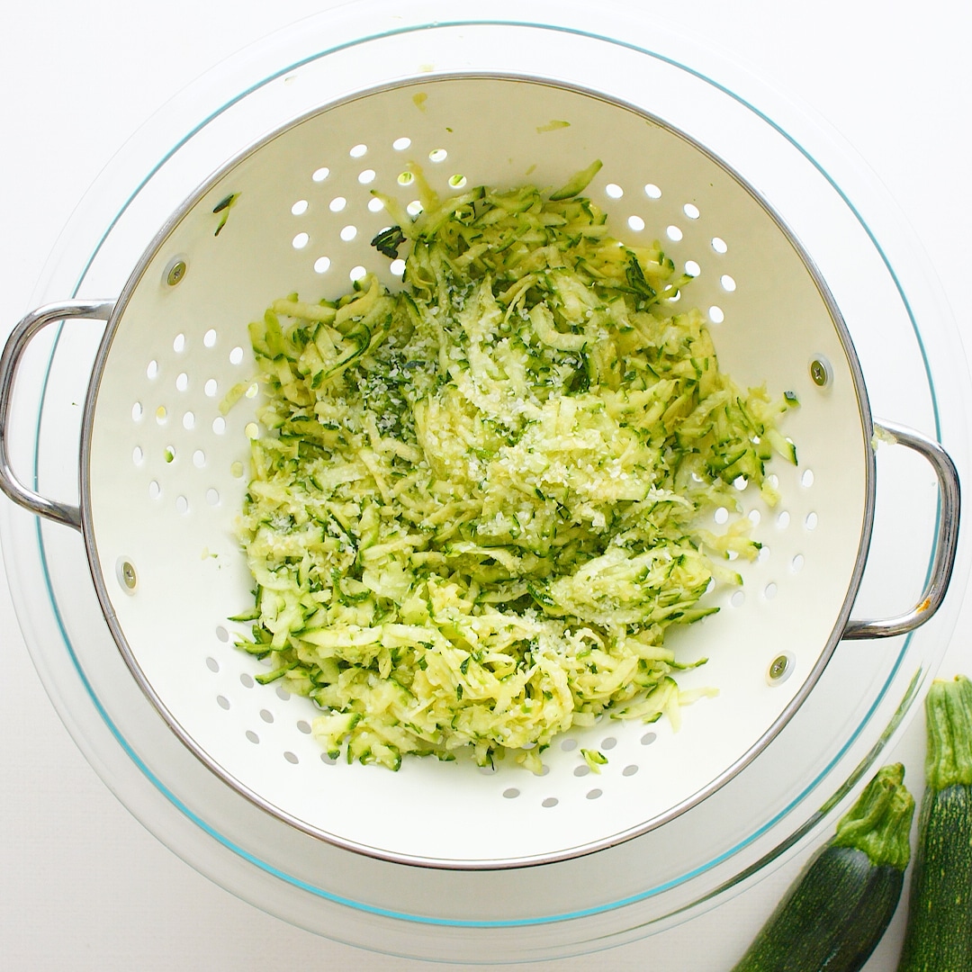 Shredded salted zucchini in a colander inside a large glass bowl