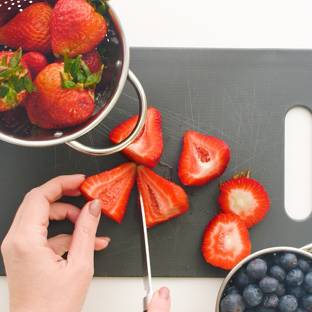 Hand holding sliced strawberry. Cutting strawberries on a cutting board. There is a strainer with strawberries and a bowl of blueberries.
