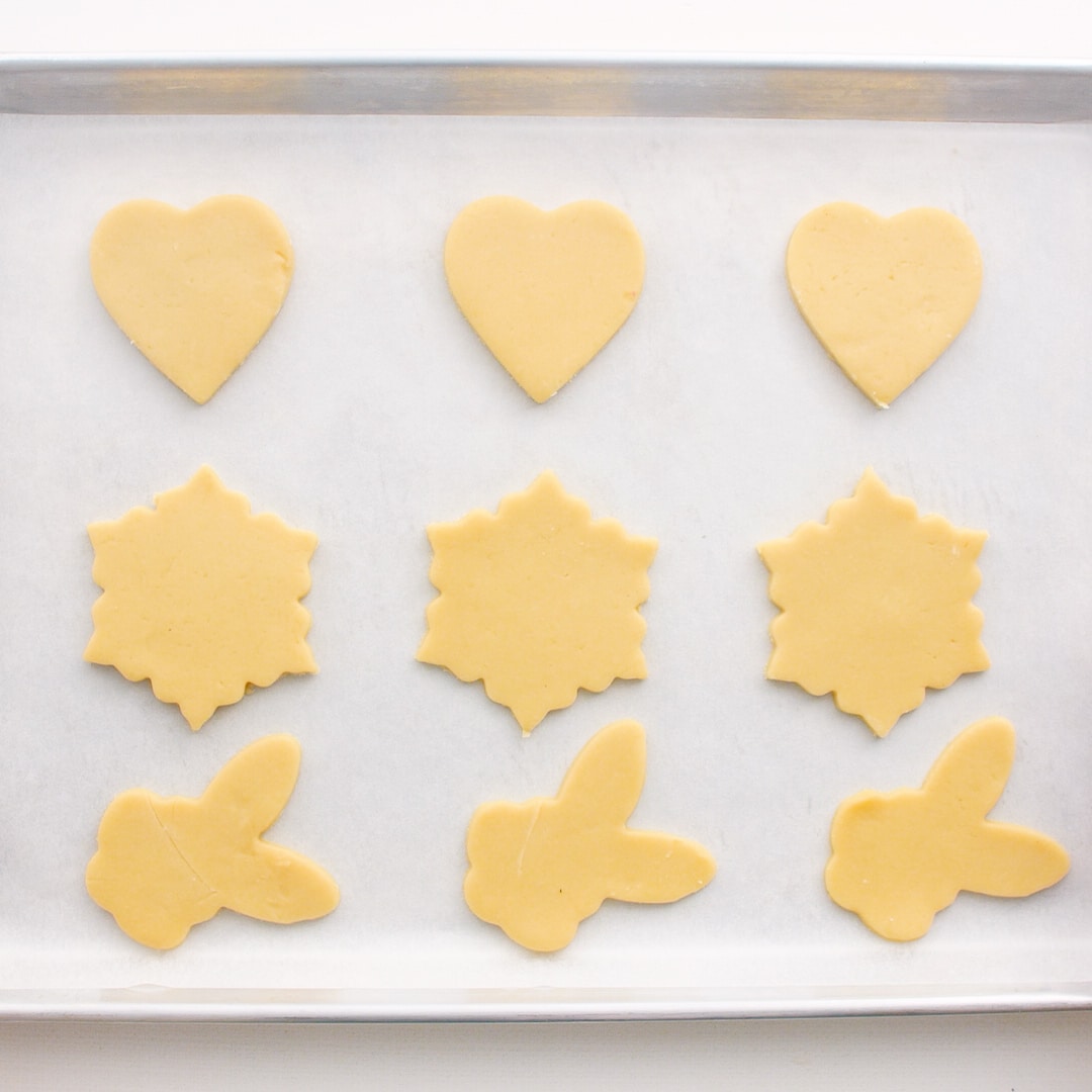 Rows of unbaked sugar cookies on a parchment paper lined baking sheet. The top row is heart shaped, the second row are snow flakes, and the bottom are bunny shaped.