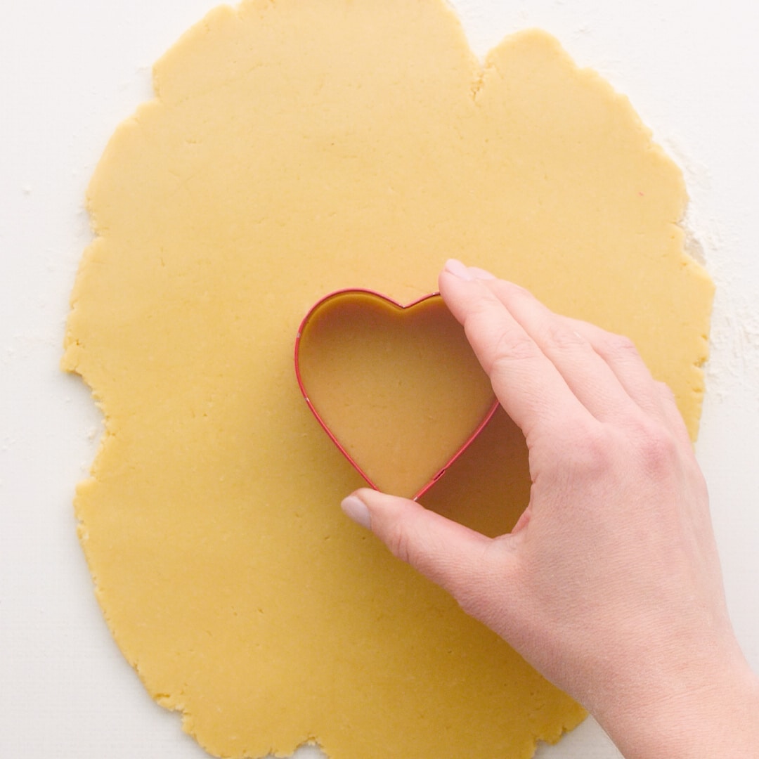 Rolled sugar cookie dough with a hand placing a heart shaped cookie cutter on the dough