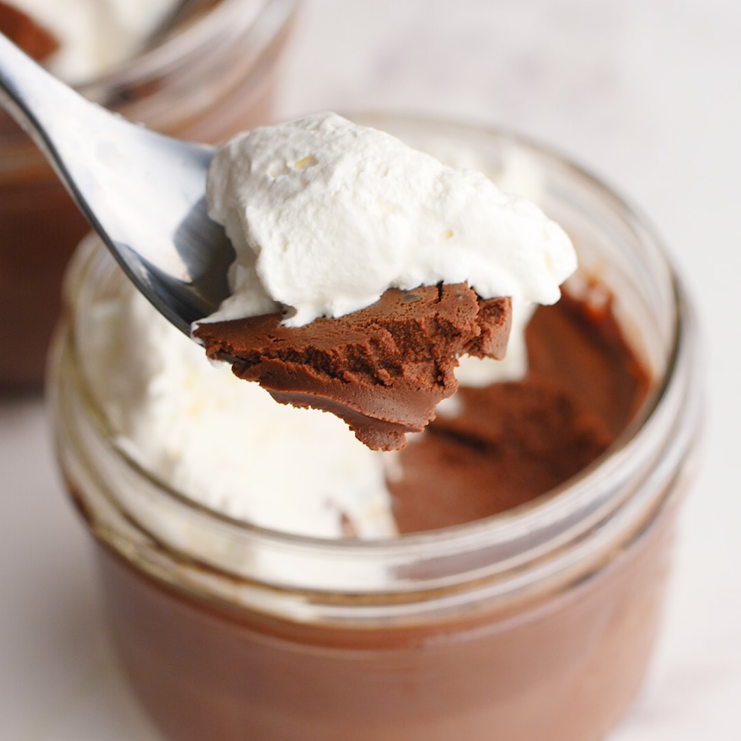 Up close view of spoonful of homemade chocolate pudding with homemade whipped cream. Mason jar full of pudding in the background