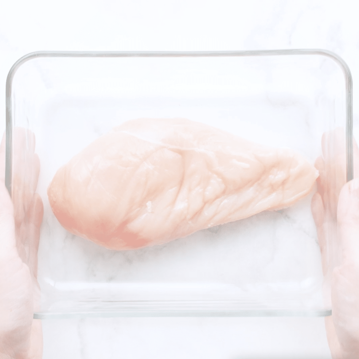 Hands holding a glass baking dish with raw chicken.