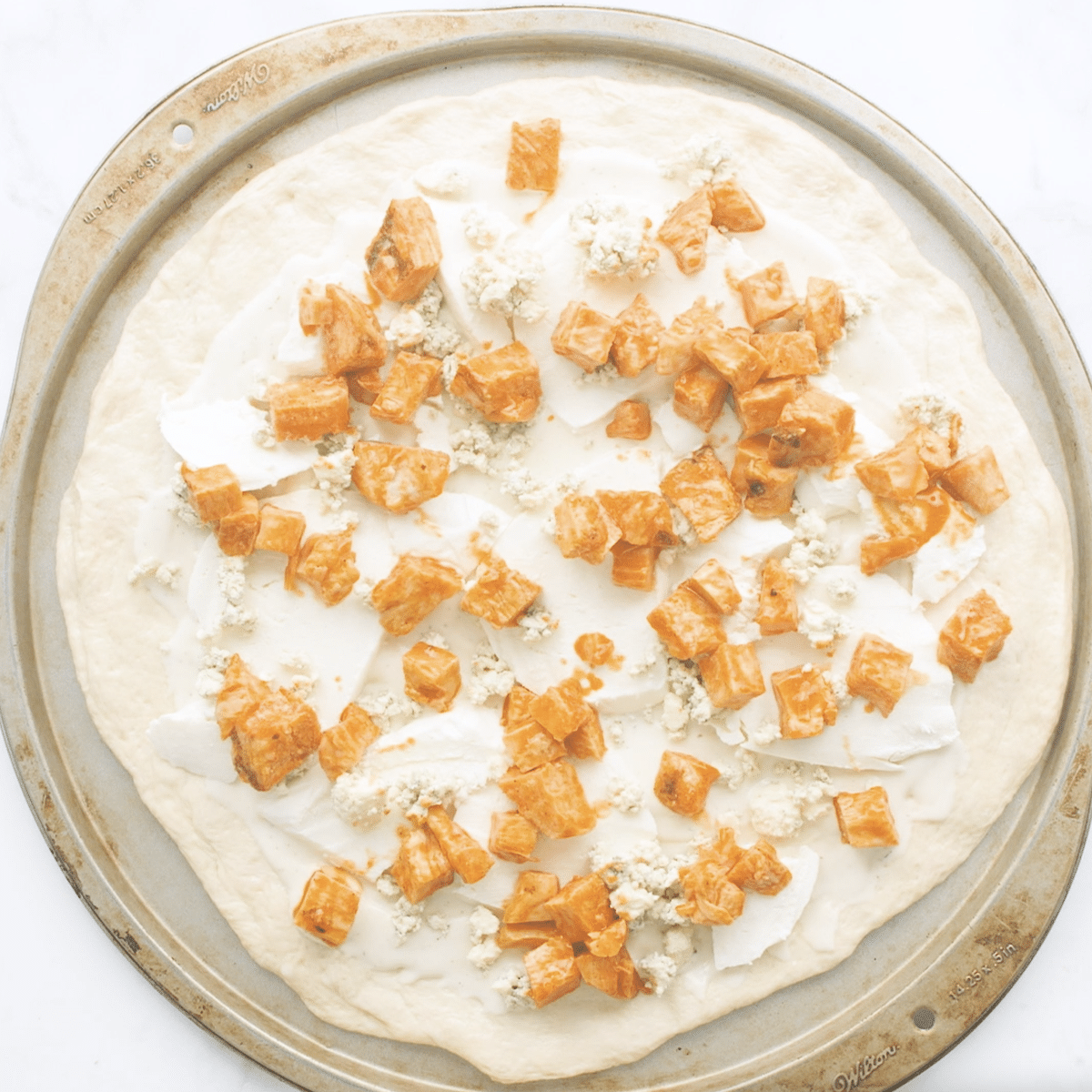 Raw pizza dough topped with buffalo chicken on a pizza stone.