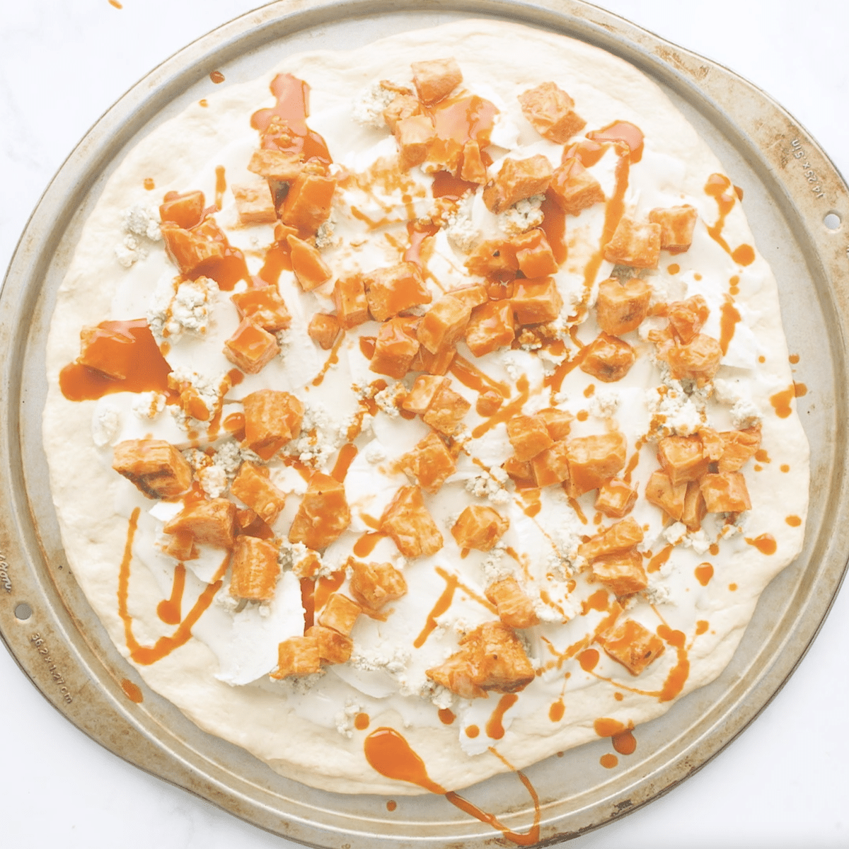 Raw pizza dough topped with buffalo chicken and hot sauce on a pizza stone.