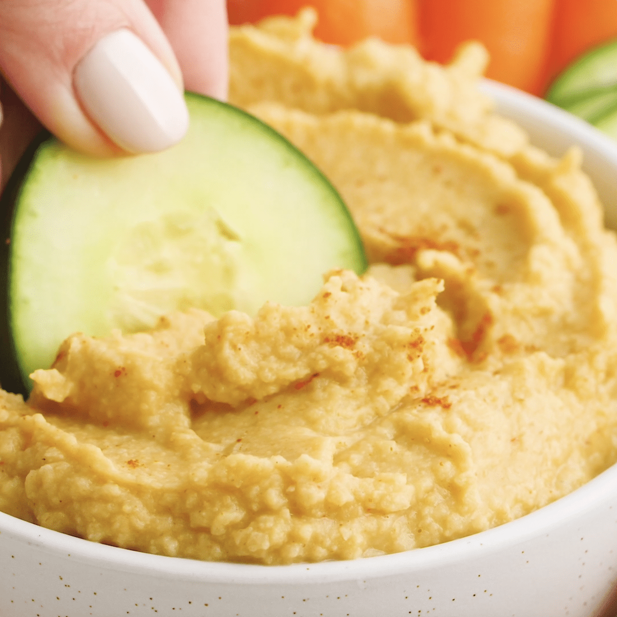 Hand holding sliced cucumber and dipping it into a bowl of homemade hummus
