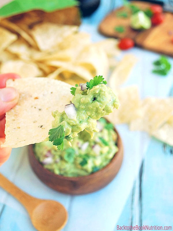 Hand holding a tortilla chip with homemade guacamole on it. Wood bowl with guacamole, wood spoon, and tortilla chips in the background.