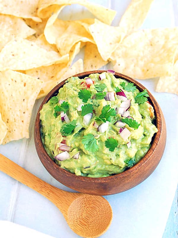Wood bowl of homemade guacamole with tortilla chips and a wood spoon nearby.