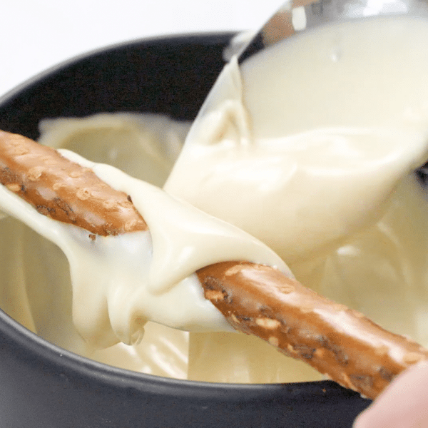 Spoon pouring melted white chocolate onto a pretzel rod. There is a bowl of melted white chocolate underneath.