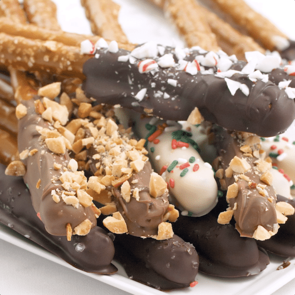 Four different variations of chocolate covered pretzels on a white plate: milk chocolate with crushed nuts, dark chocolate, white chocolate with holiday sprinkles, and dark chocolate with crushed candy cane bits.