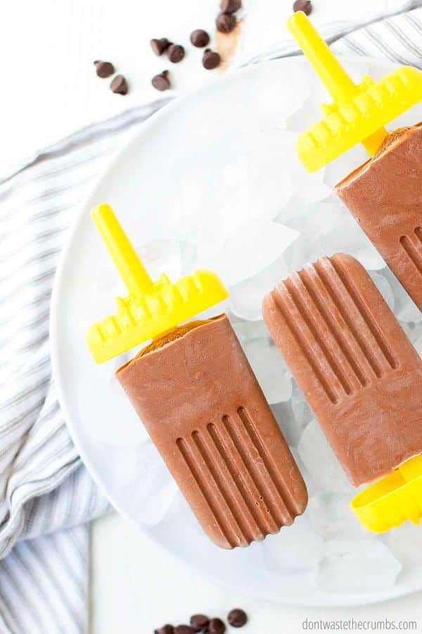 Homemade fudge pops lying in a bowl of ice, on top of a dish towel, with some chocolate chips scattered nearby