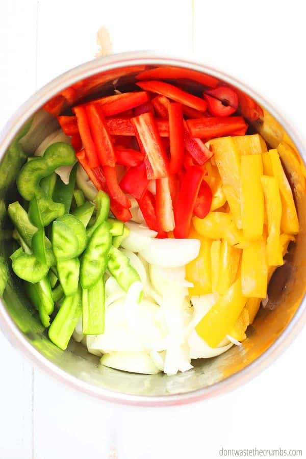 Raw sliced red, green, and yellow bell peppers, in strips, with sliced raw onions inside of an Instant Pot - ready to be cooked as fajita veggies.