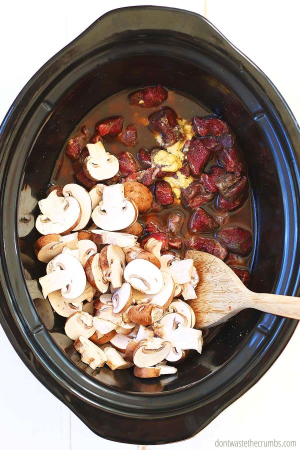 Sliced mushrooms, beef broth, Worcestershire sauce, mustard, and spices over raw cut beef in the crock pot. There is a wooden spoon stirring the ingredients.