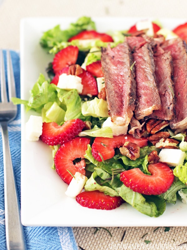 Strawberry salad recipe topped with sliced steak on a white plate with a fork next to it.