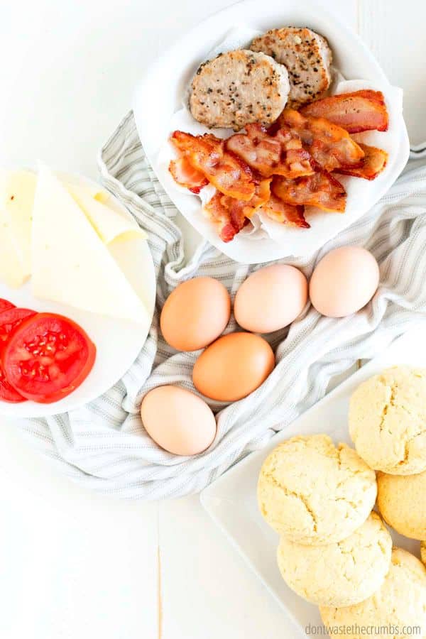 Ingredients for breakfast sandwiches: sausage, bacon, cheese, sliced tomato, eggs, and biscuits.