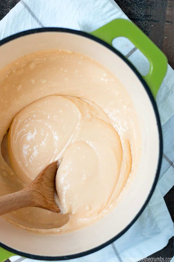 Freshly cooked cheese sauce in a green Dutch oven being stirred with a wooden spoon. 