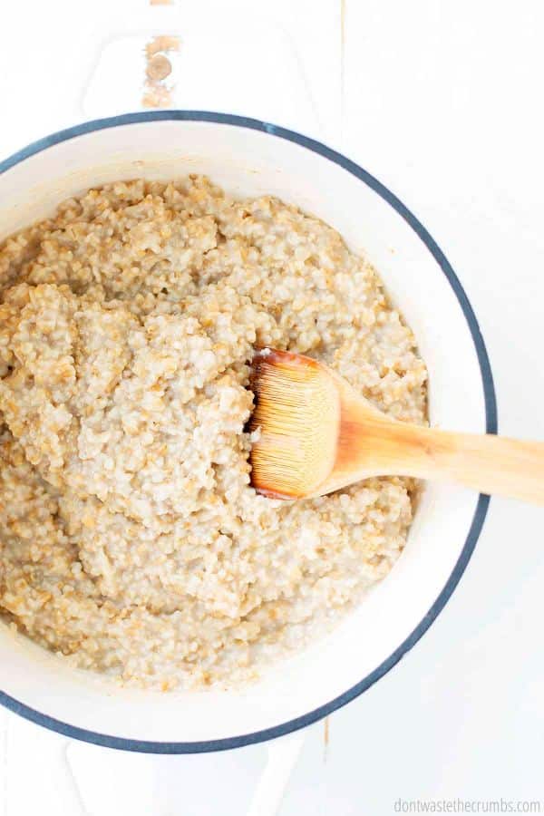 A dutch oven is filled with cooked steel cut oats. A wooden serving spoon is inserted into the oatmeal.