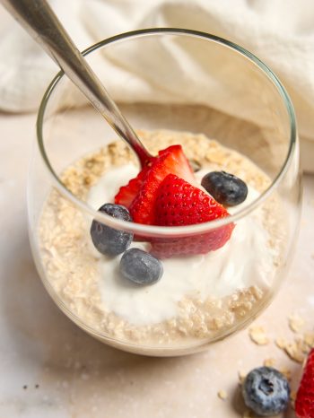 A glass of overnight oats with a spoon sticking out, topped with yogurt, sliced strawberries, and blueberries.