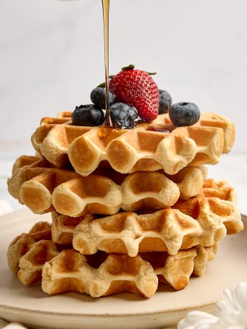 A stack of waffles with protein, topped with fresh blueberries and strawberries, with syrup pouring over the top from a small pitcher
